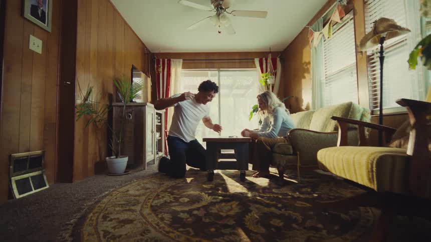A man and a woman are playing a board or card game in a cozy woodpaneled living room decorated in a vintage style the man kneeling on the rug throws his hands up in a gesture of success or frustration while the woman sitting on the couch covers he