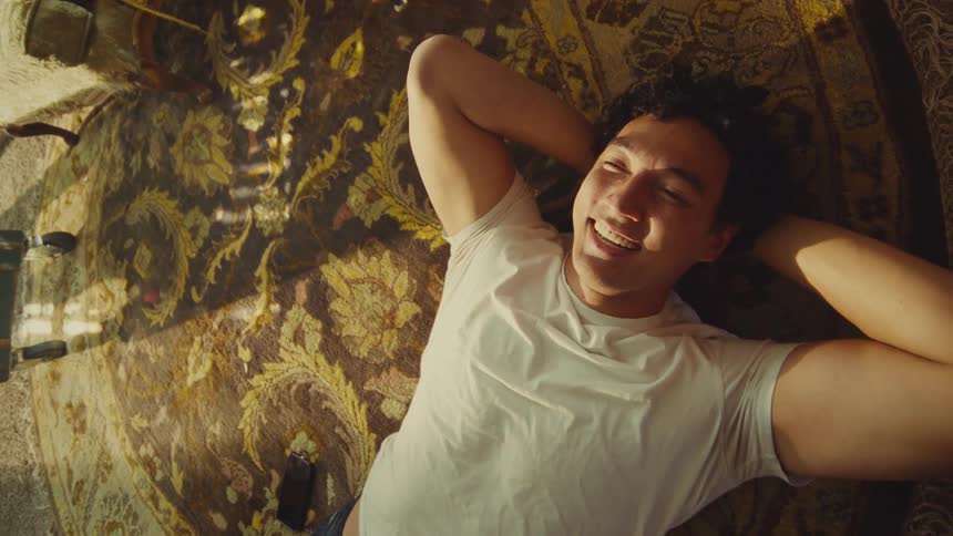 A man with curly dark hair wearing a white tshirt lies on his back on an ornate patterned rug initially stretching his arms behind his head and smiling then bringing a smartphone into the frame and engaging with the device
