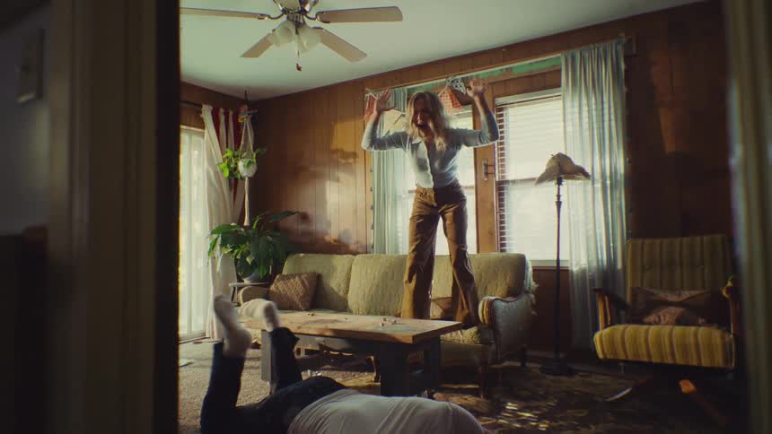 A woman triumphantly jumps on a couch with her hands raised as a man collapses on the floor in a sunlit retro living room with wood paneling and a shag rug