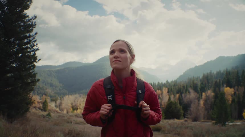 Young adult woman wearing a red jacket and carrying a backpack walking around a landscape with a forest and mountains on the background outdoors during day time