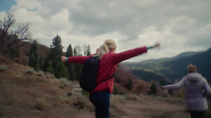 Two people hike mountain trail with autumn trees and snowy peaks under partly cloudy sky