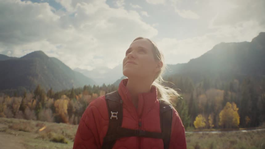 A young woman in a red jacket carrying a backpack hikes through the woods on a sunny day