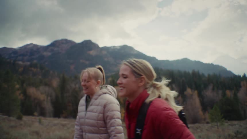 Two people in puffer jackets walk through mountainous terrain surrounded by pines and peaks under cloudy sky