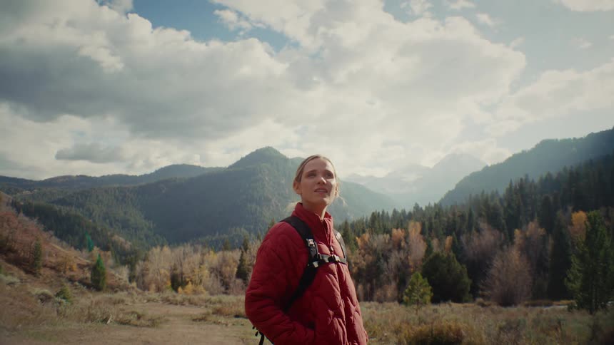 A female hiker in a red jacket stands in a mountain valley gazing around at the scenery as sunlight flares behind her