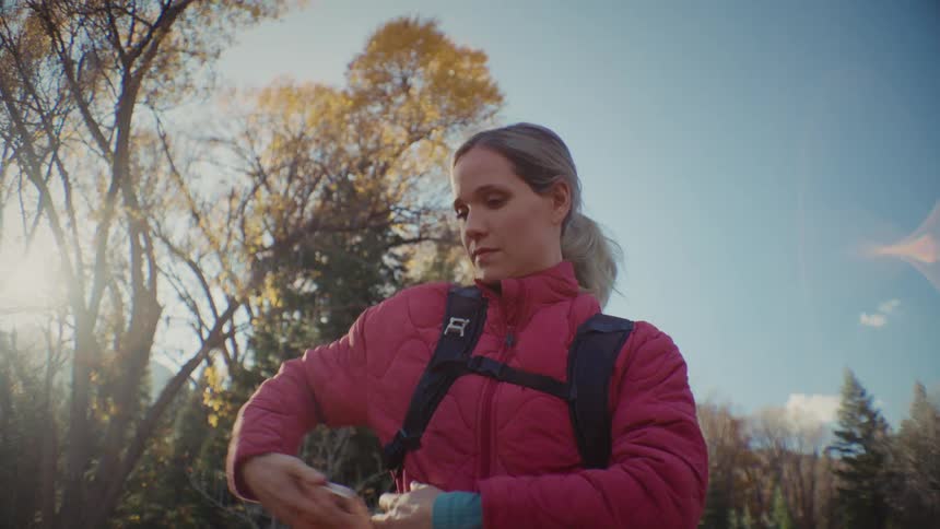 Young woman wearing a red jacket and carrying a backpack using a smartphone before putting it on her pocket and moving forward next to a forest outdoors during day time