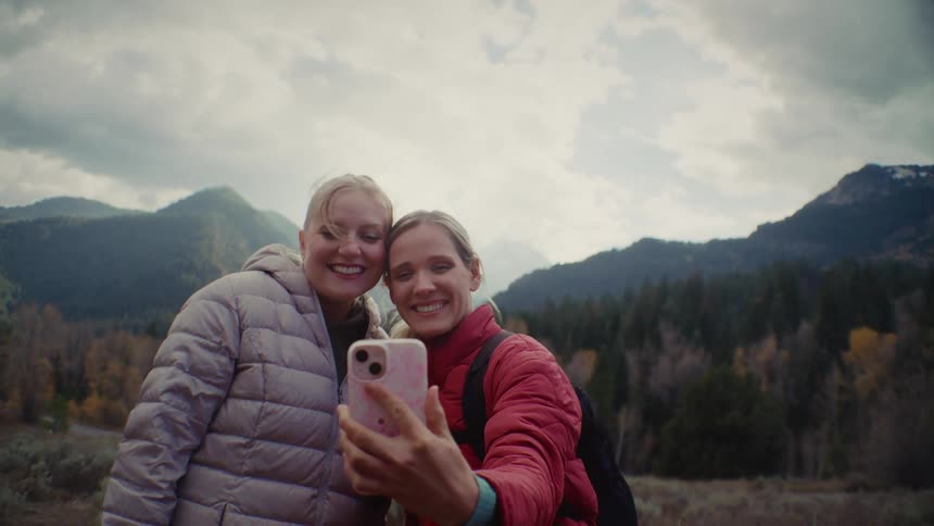 Two smiling women wearing puffer jackets take a selfie with a pink smartphone against a scenic mountainous landscape under an overcast sky