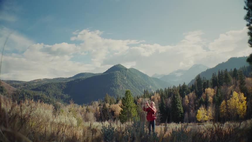 A woman in a red jacket stands in a scenic mountainous valley covered in evergreens and autumn trees raising her hand to take a photo of the view