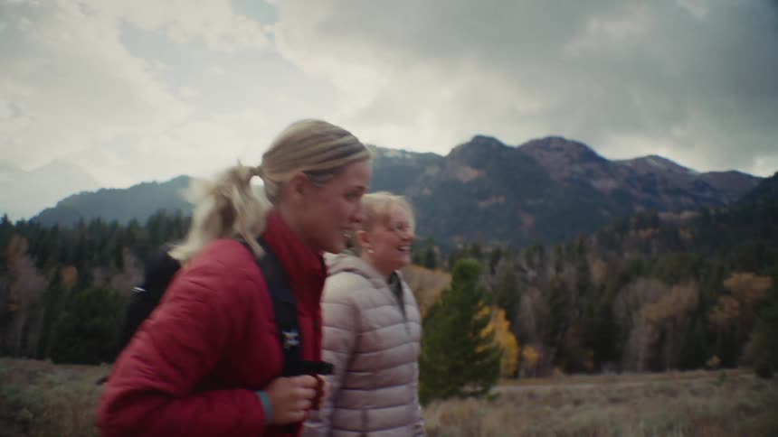 Two smiling women hike along a trail in a mountain landscape during autumn
