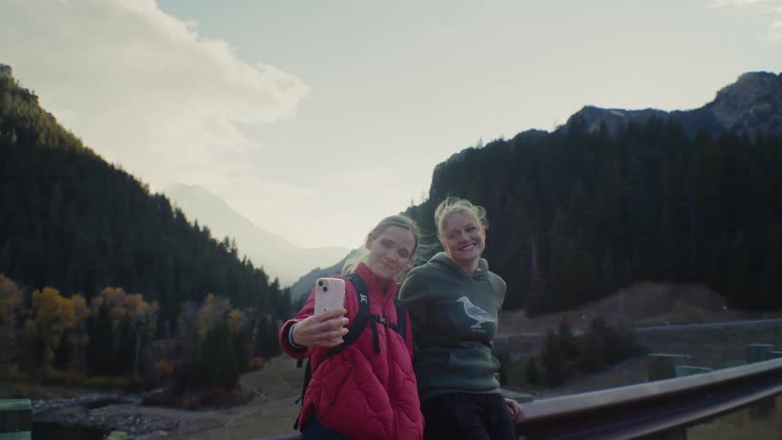 Two young women dressed in outdoor gear stand near a roadside guardrail in a deep mountain valley and smile while one of them holds a mobile phone to capture a selfie of themselves with the forested mountain backdrop