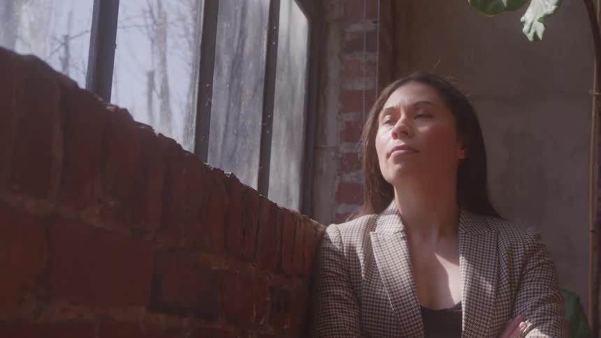 Smiling adult woman wearing a blazer standing against a brick wall indoors next to a window during day time