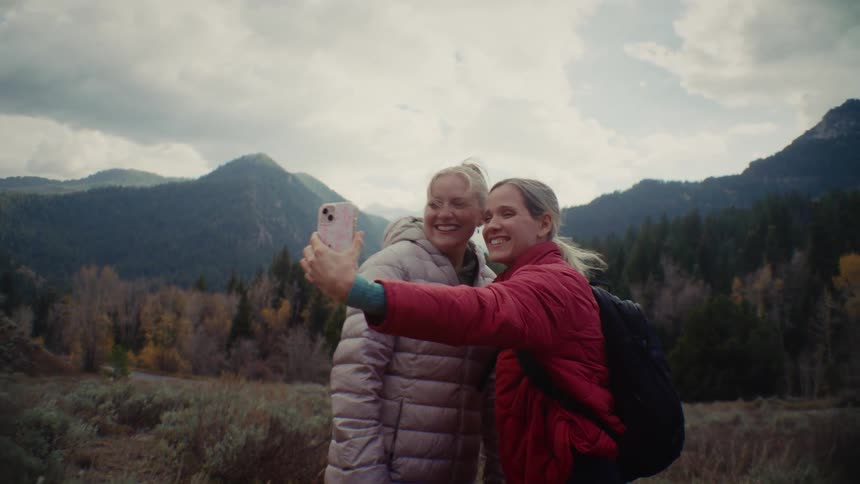 Two women pose for a selfie with mountains and a wooded valley behind them
