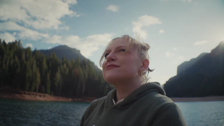 A young woman is outdoors on a sunny day with a lake mountains and forest forming the background