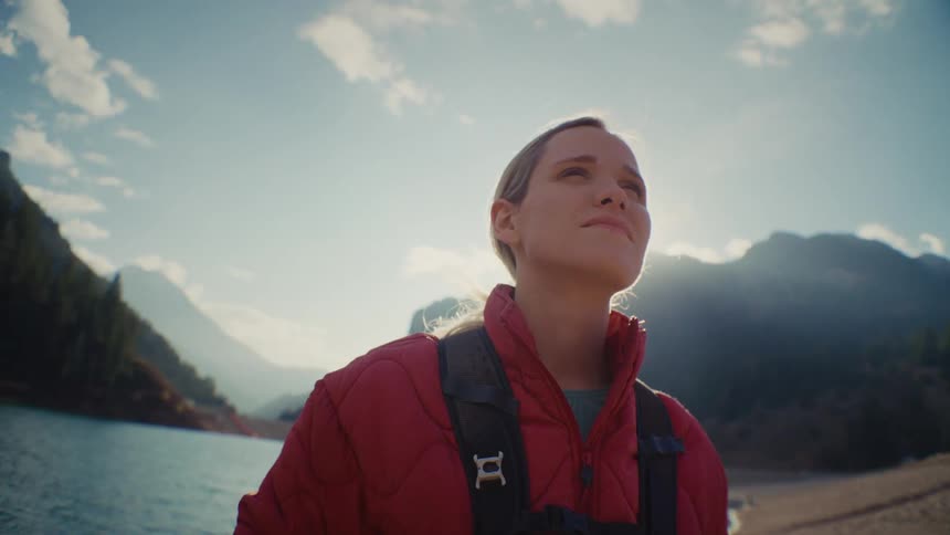 Woman with a backpack looks at the sunlit mountain landscape by a lake