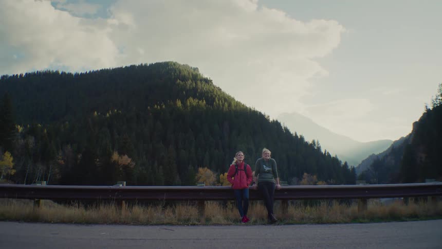 Two friends stand smiling by a guardrail on the side of a mountain road with a forested hillside and deep valley behind them under a bright sky