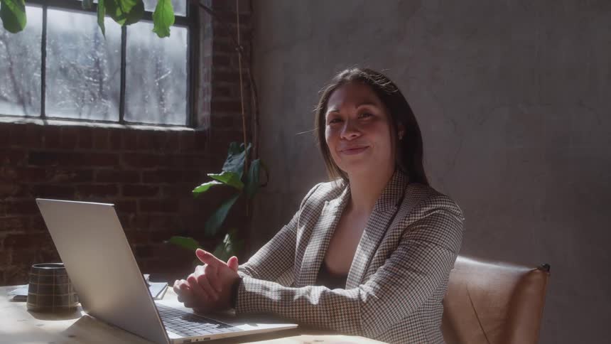 Woman in checkered blazer smiles gently at laptop in warmly lit cozy workspace