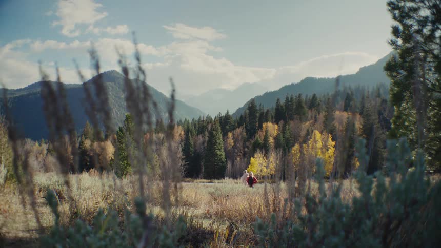 A couple runs through a golden autumn field framed by tall grasses with a dense forest and rugged mountains rising in the background