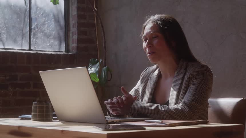A woman wearing a blazer sits at a wooden table and smiles while talking animatedly during a video call on her laptop in a sunlit loft space
