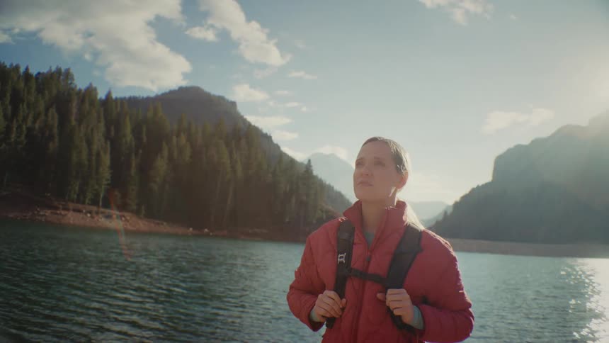 A woman wearing a red jacket and backpack smiles and looks up at the sky while standing by a clear mountain lake surrounded by dense evergreen forests