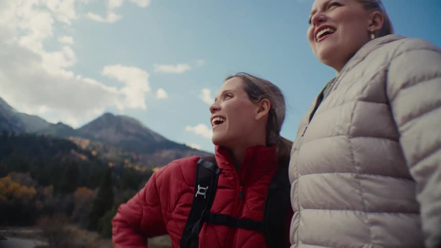 Two laughing women in puffer jackets stand together and look around in a scenic mountain landscape