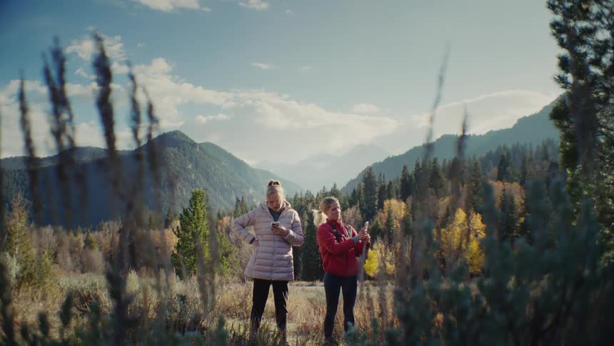 Two women dressed for cold weather stand in an open grassy wilderness area framed by mountains and evergreen trees with yellow autumn leaves as one of them holds her phone up to search for a signal