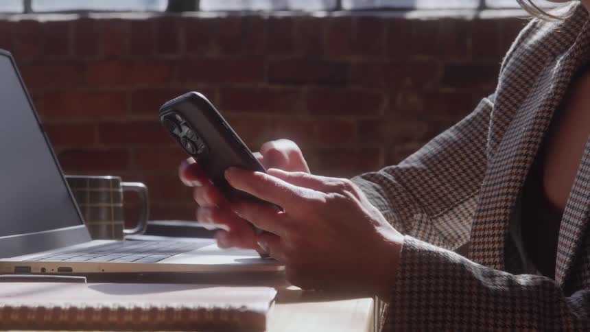Woman sits at a desk with a laptop computer as she holds her phone texting with her thumbs