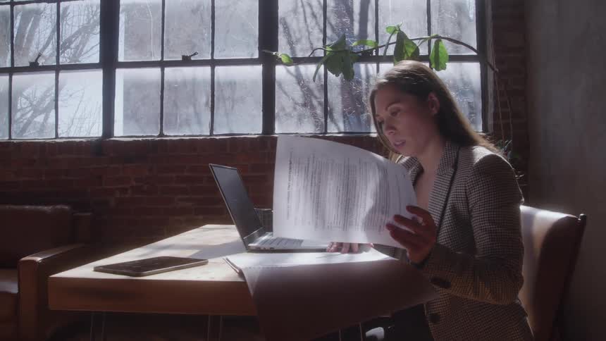 Woman seated at a desk rests her head on her hand while working at a laptop in sunlit surroundings
