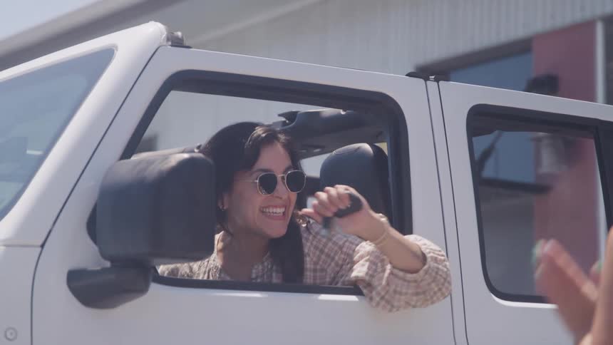 A smiling woman wearing sunglasses and a plaid shirt sits in the drivers seat of a white offroad vehicle with the top down leaning out the window to receive a key or small object from another person offcamera