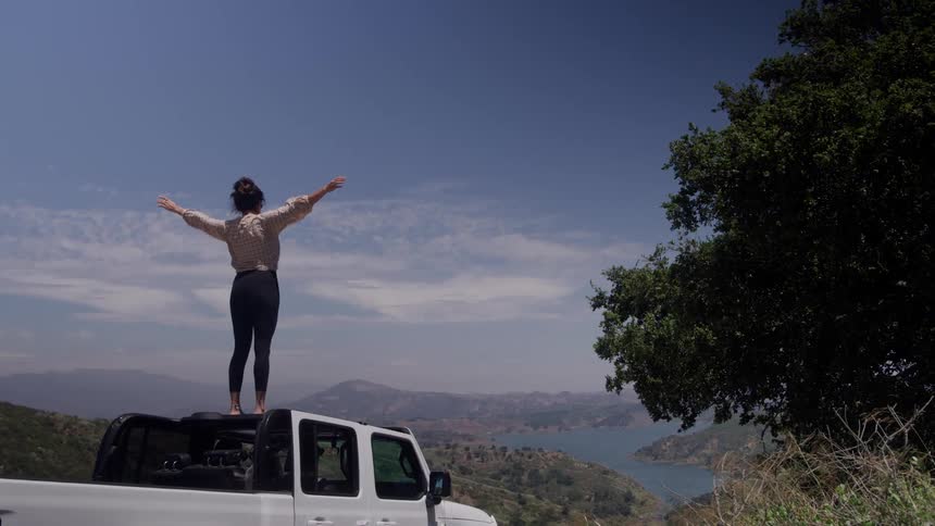 Woman stands on the roof of her truck and raises her arms in the air