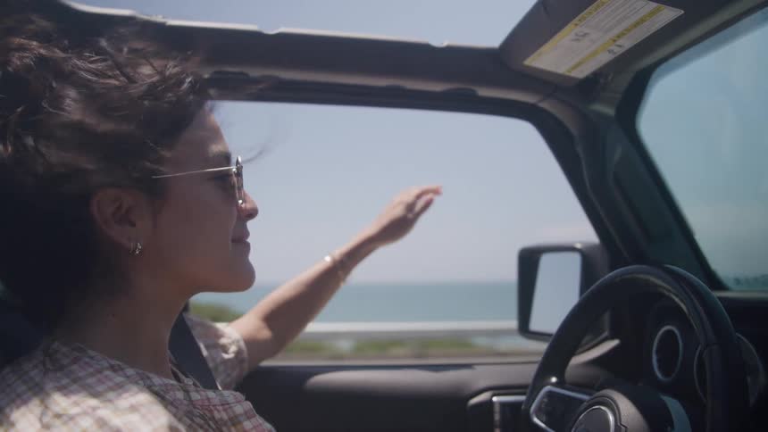 A woman driving a convertible along a coastal road smiles as she lets her hand glide through the wind