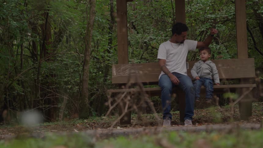 A man and his small son sit on a wooden bench inside a covered shelter surrounded by a dense green forest enjoying a peaceful time outdoors