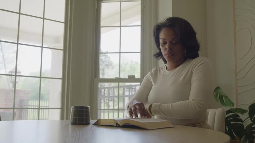 A middleaged black woman sits behind a dining table at midday opening her bible and leaning forward to read from it