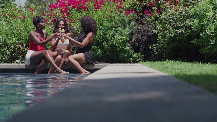 Three women sitting by the poolside toasting amid a flowering garden