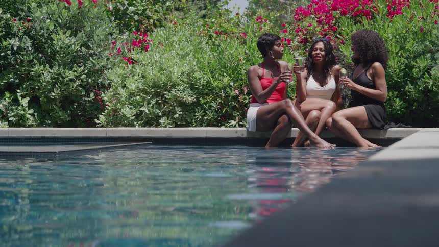 Three young women in swim attire sit on the edge of a swimming pool holding champagne glasses and talking in front of bright red flowers and green foliage
