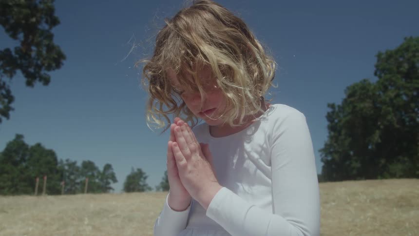 This video shows a young girl outdoors on a sunny day she has blonde curly hair and is wearing a longsleeved white topshe is seen with her eyes closed holding her hands together in a prayer or meditative pose against the backdrop of a clear blue sk