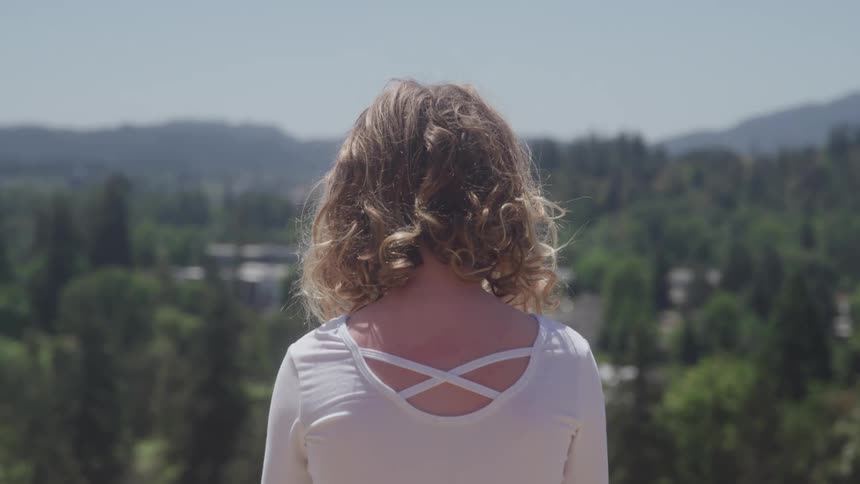 This video features a medium shot of a woman with curly light brown hair viewed from behind standing on a vantage point she is wearing a white top with a crisscross detail on the back and is looking out over a lush green landscape of rolling hills an