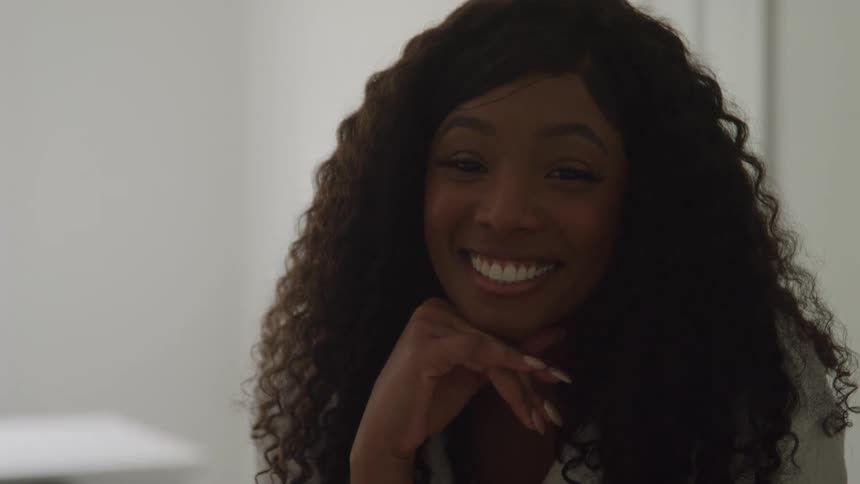 A smiling woman with long dark curly hair resting her chin on her hand as she looks offcamera with a bright engaged expression in a softly lit interior space