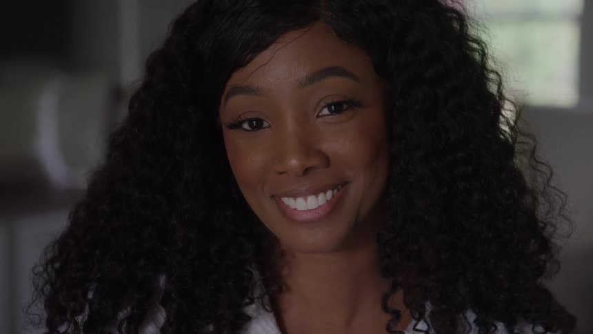 The video is a closeup of a woman with long dark curly hairshe initially looks to the side then slowly turns her gaze toward the camera and offers a warm genuine smile