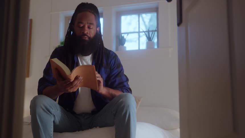 Man sitting on the bed reading a book in a bright bedroom