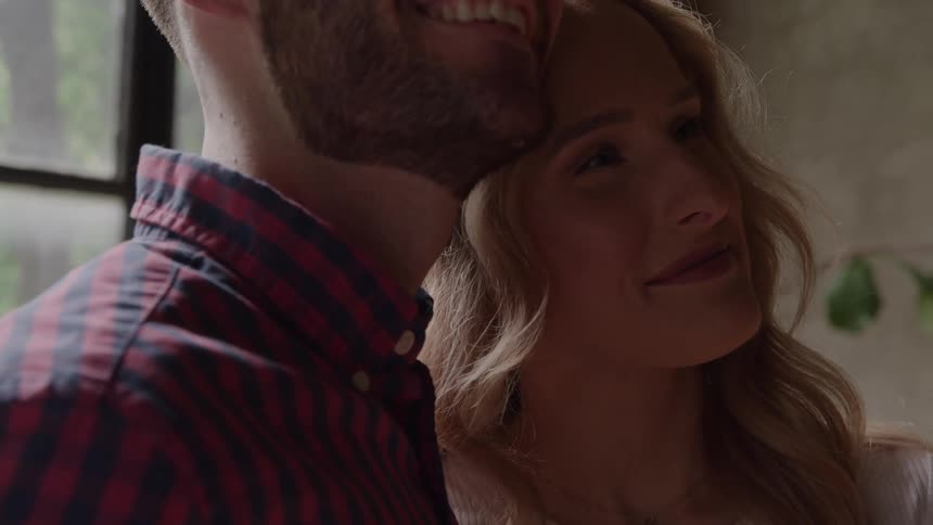 A closeup shot of a man and a woman standing close together with the mans face partially visible as he smiles and the woman leaning into him with her eyes closed showing an expression of serene happiness
