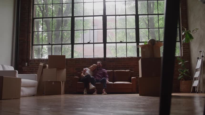 A couple sits together on a couch surrounded by moving boxes in an empty loft apartment with large industrial windows