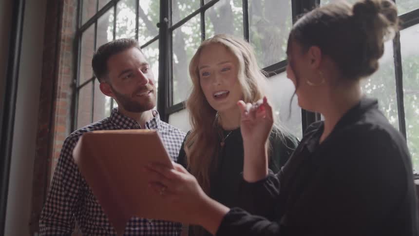 The video shows three people standing together near a large industrialstyle window engaged in a discussion a woman is holding a clipboard or tablet and gesturing as she speaks to the couple who are smiling and reacting positively to what she is prese
