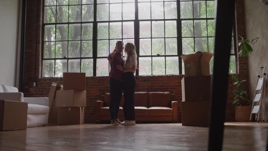 A couple moves into a spacious loft apartment carrying boxes toward a large window overlooking the exterior foliage they stop to embrace and admire the high ceilings and vast open space of their new home surrounded by moving boxes and their first piece