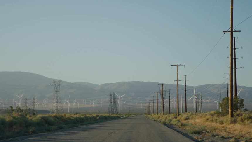 Utility poles and turbines spinning on a wind farm along a rural road with mountains in the distance