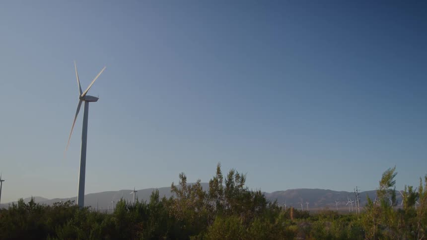 A static wide shot captures a large white wind turbine spinning its blades above desert brush with mountains visible in the distance under a clear blue sky