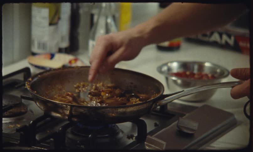 A persons hands stir a savory mixture cooking in a stainless steel pan over a gas stovetop in a kitchen with various items in the background