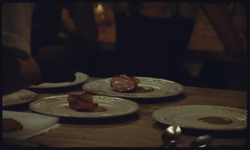 A persons hands carving slices of cooked red meat on a white cutting board then plating individual portions onto several white plates each already containing a smear of brown sauce in a restaurant kitchen