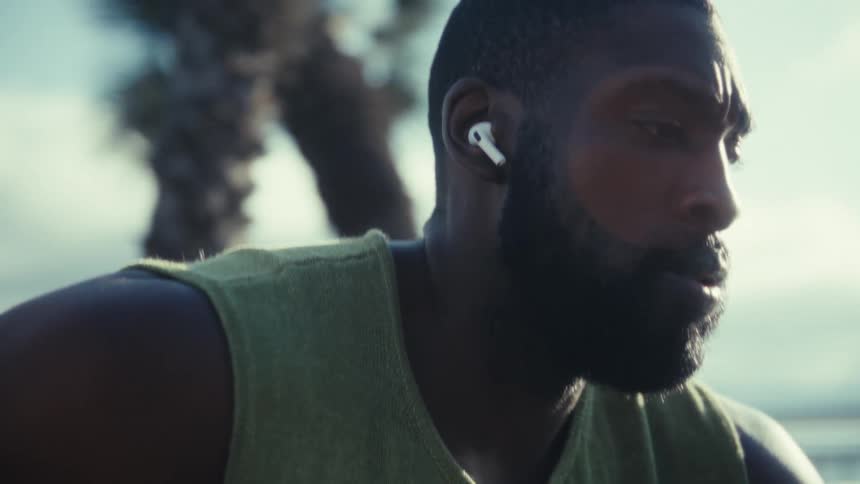 The video is a closeup of a man outdoors sweating wearing a tank top and a wireless earbud in his right earhe is initially silhouetted against bright sunlight then the camera adjusts to reveal his face as he breathes heavily suggesting he is paus