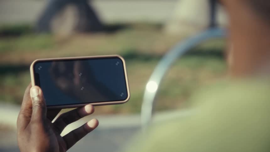 A close view of a dark skinned hand holding a modern smartphone with a blank screen outdoors in bright sunlight
