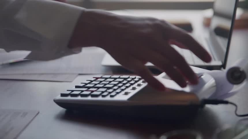 Hands type on a desktop calculator beside a laptop