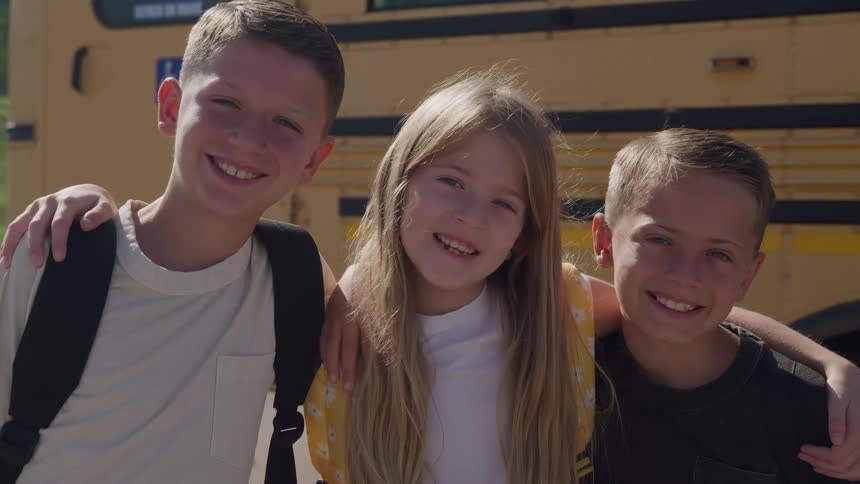 Three smiling children two boys and one girl stand together with their arms around each other in front of a yellow school bus on a sunny day
